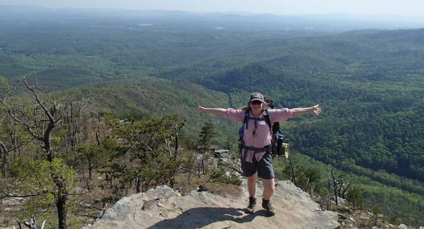 A person wearing a backpack stands on a rocky overlook with their arms spread wide. Below them, the blue ridge mountains stretch for miles. 